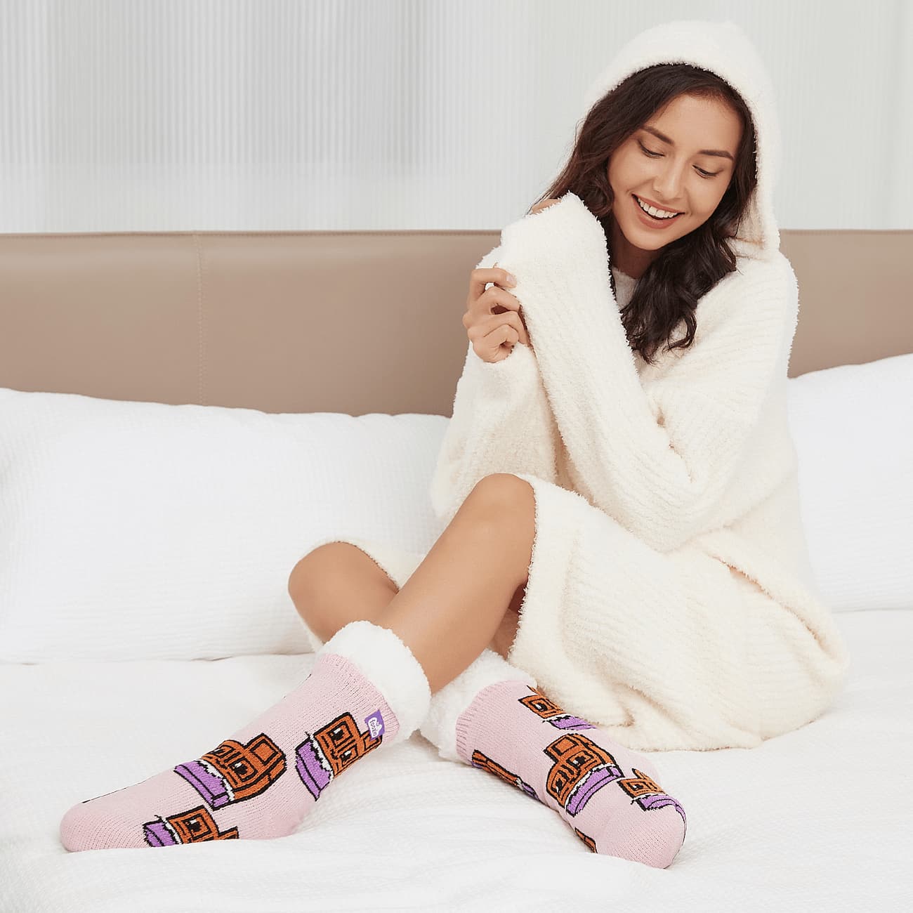 The Foozie™ Chocolate slipper socks, worn by a woman with her hood up, sitting on a white bed