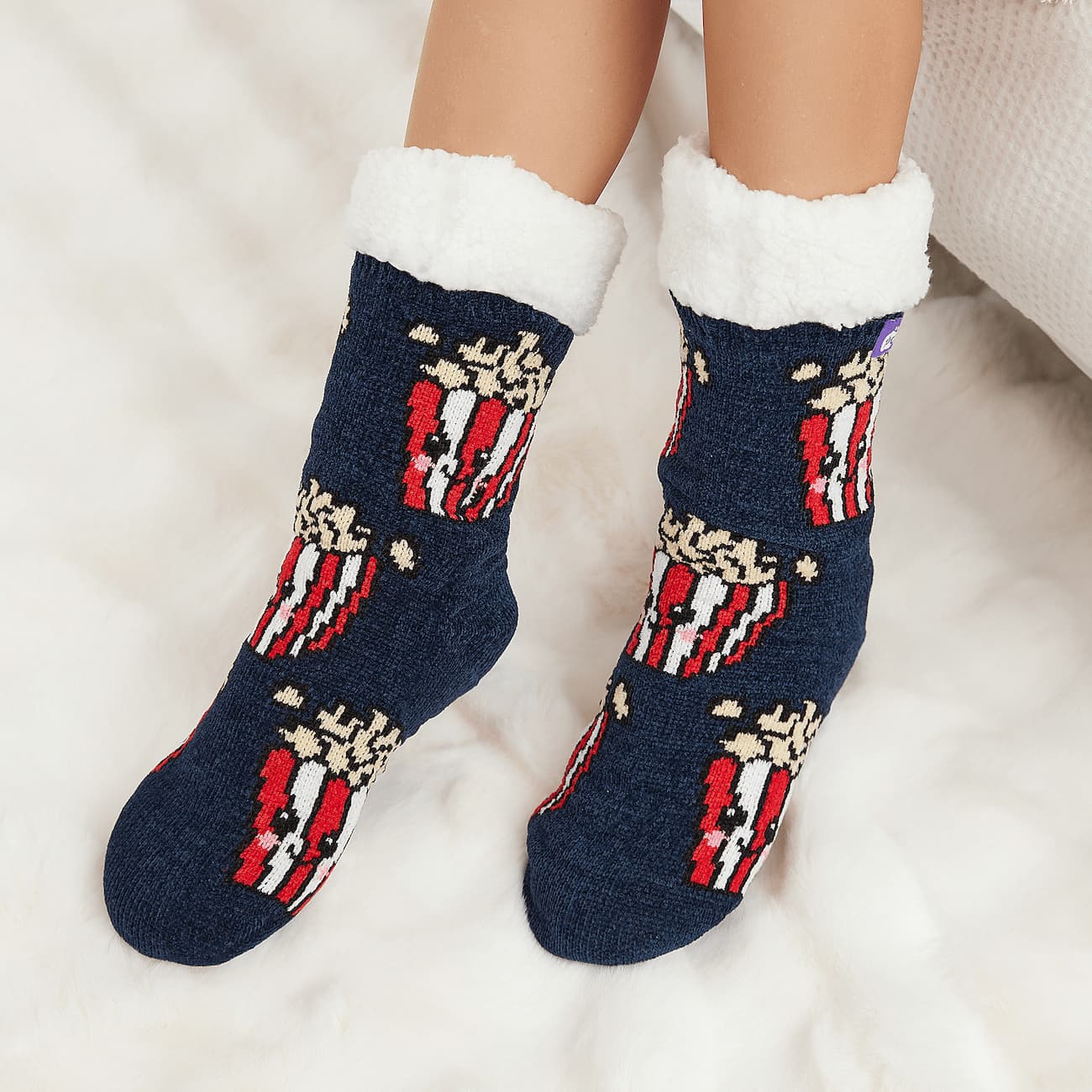 The Foozie™ Popcorn slipper socks, smiling popcorn box print on navy, worn on a fluffy white rug